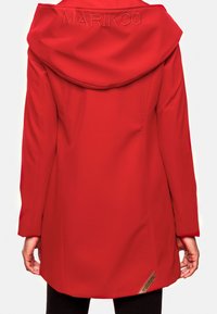 Chaqueta roja con capucha de tela suave, con una capucha grande y caída, y una etiqueta triangular de la marca en el dobladillo. Cierre no visible.