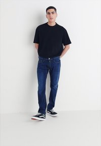 Svart kortärmad t-shirt, blå denimjeans och svarta sneakers med vita sulor. Modellen står mot en enkel vit bakgrund.