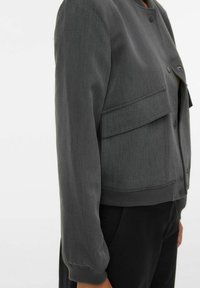 Szara krótka kurtka wykonana z gładkiej tkaniny, z okrągłym kołnierzem, zapięciem na guziki oraz bocznymi kieszeniami. Rękawy mają marszczone mankiety.