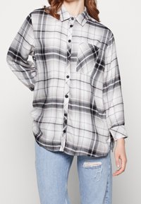 Camicia a quadri bianco e nero con maniche lunghe, chiusura a bottoni, taschino sul petto e una vestibilità ampia e rilassata. Il materiale sembra morbido e con una texture.