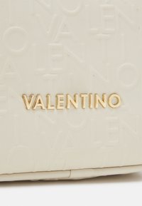 Krämfärgad läderväska med präglad logotyp och en guldtonad "VALENTINO"-logotyp som är tydligt placerad i mitten.