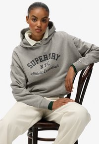 Sudadera gris con capucha que lleva el texto "SUPERDRY N.Y.C Athletic Essentials". Presenta un bolsillo tipo canguro, puños acanalados y un cuello verde en capas. Pantalones crema.