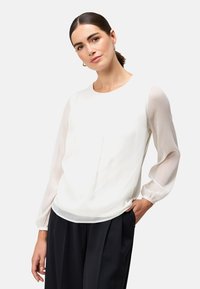 Weiße Bluse mit transparenten, langen, sanft gepufften Ärmeln; strukturierter Körper mit dezenten Streifen; runder Ausschnitt und lässige Passform. Kombiniert mit schwarzen Hosen.