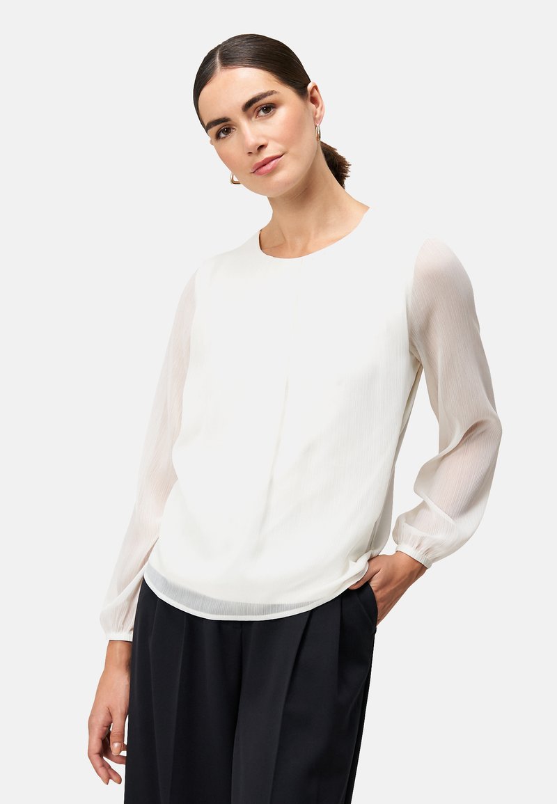 Weiße Bluse mit transparenten, langen, sanft gepufften Ärmeln; strukturierter Körper mit dezenten Streifen; runder Ausschnitt und lässige Passform. Kombiniert mit schwarzen Hosen.