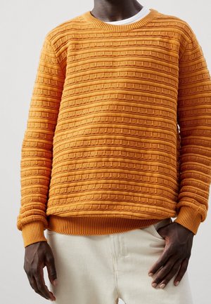 Personne portant un pull en maille orange texturé par-dessus une chemise blanche, combiné avec un pantalon beige clair, debout devant un fond uni.
