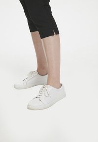 Zapatillas de malla blancas con cordones blancos, con puntera redonda y suela de goma, combinadas con pantalones cortos negros. Textura sutil visible en los zapatos.