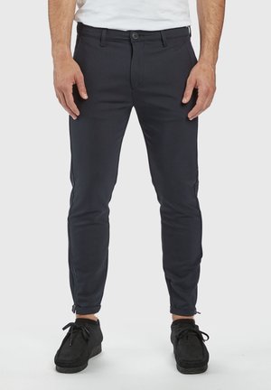Hombre con pantalones negros ajustados con cremalleras en los tobillos y zapatos negros con cordones, de pie contra un fondo claro y liso.