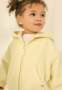 Sweat à capuche zippé jaune avec une grande poche avant, une capuche et une texture douce. Présente une petite étiquette à l'ourlet avec un détail de logo.