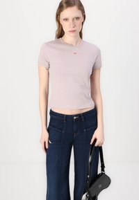 T-shirt corta di colore viola chiaro con maniche corte e piccolo logo rosso sul petto, abbinata a jeans a gamba larga di denim scuro e una borsa nera.