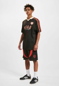 Costum sport negru format dintr-un tricou lejer cu un logo roșu și accente, completat de o pereche de pantaloni scurți negri cu panouri laterale roșii și logo.