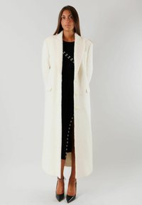 Lungo cappotto bianco di lana con colletto a punta, chiusura con bottoni, due tasche frontali, abbinato a un vestito nero a costine e scarpe con tacco a punta nere.