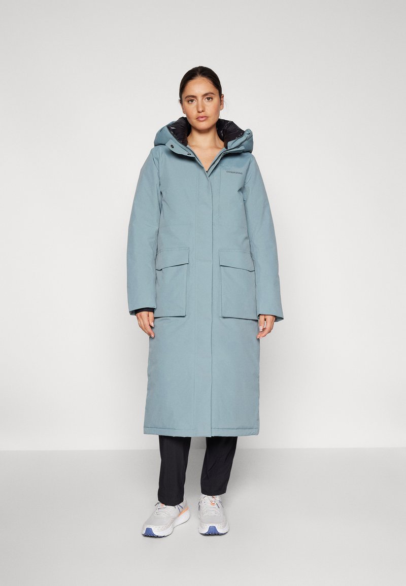 Cappotto impermeabile lungo di colore blu chiaro con cappuccio. Presenta due tasche frontali, una chiusura con zip ed è indossato sopra pantaloni scuri e sneakers.