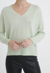 Pull vert clair à col en V avec poignets et ourlet côtelés, fabriqué en matière douce. Présente une texture subtile et un petit logo métallique en accent.