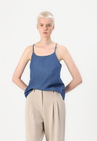 Denim blue sleeveless top with thin shoulder straps, a straight neckline, and a relaxed fit. Worn with beige pleated trousers.