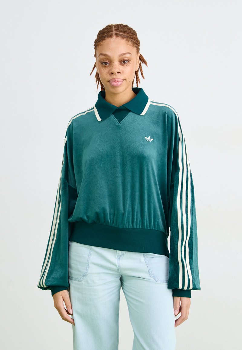 Jong persoon met vlecht haar, gekleed in een tealkleurig Adidas t-shirt met lange mouwen en witte strepen, met lichtblauwe jeans, staande tegen een effen achtergrond.