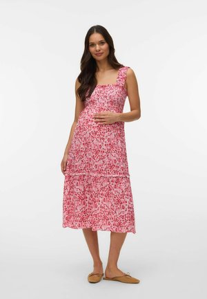 Femme aux longs cheveux bruns portant une robe midi fleurie rose sans manches, tenant son ventre, debout en ballerines beige sur un fond blanc.