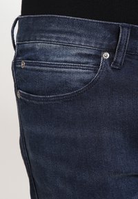 Vaqueros de mezclilla azul oscuro con una vista en primer plano del bolsillo, tela texturizada y remaches metálicos. El diseño incluye sutiles detalles de costura.