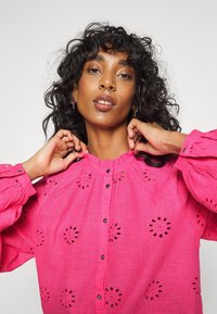 Blusa rosa, bordada, com um colarinho alto em babados, botões pretos e mangas bufantes com padrões florais recortados.
