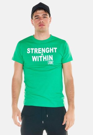 Jonge man draagt een groen t-shirt met witte tekst ""STRENGHT IS WITHIN"" en een zwarte pet, staand tegen een effen achtergrond.