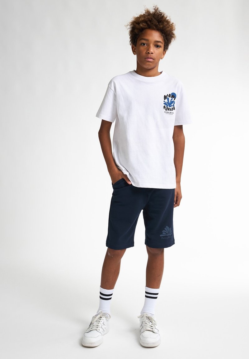 Witte katoenen t-shirt met blauwe graphic op de borst, gecombineerd met marineblauwe shorts, witte sneakers en zwartgestreepte sokken.