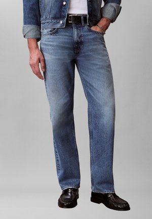 Vīrietis, kurš valkā gaiši zilu džinsa bikses, melnu jostu, melnas ādas kurpes un džinsa jaku ar uzrullētiem piedurknēm, viena roka kabatā.