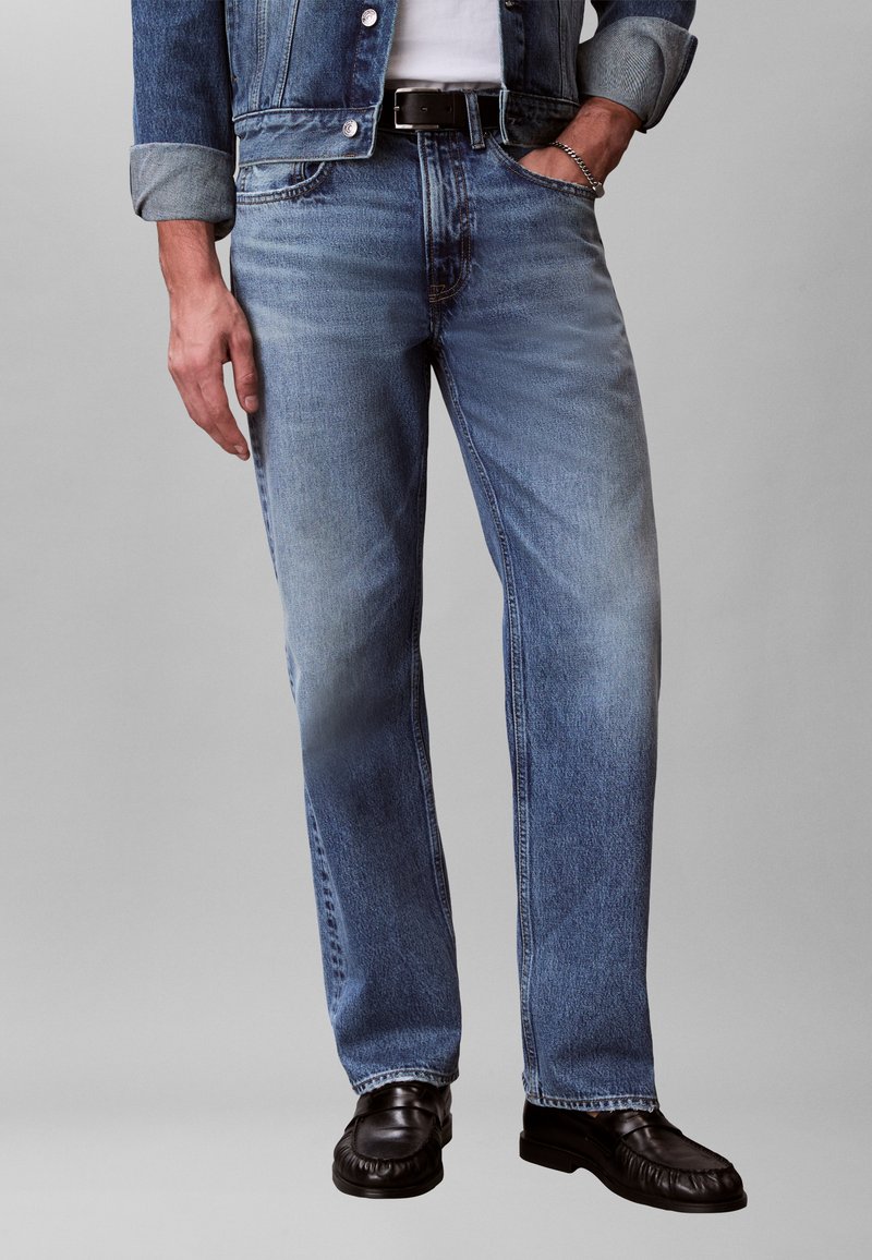 Άντρας που φοράει ανοιχτό μπλε τζιν παντελόνι, μαύρη ζώνη, μαύρα δερμάτινα παπούτσια και τζιν μπουφάν με τα μανίκια γυρισμένα, ένα χέρι στην τσέπη.