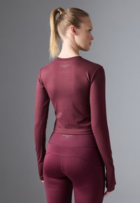 Maroon long-sleeve active top with fitted design and ribbed structured hem, paired with matching high-waisted leggings, smooth texture.