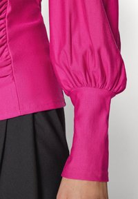 Fitted rosa topp med rynkdetaljer på sidan, puffärmar och en slät yta, tillsammans med en svart kjol.