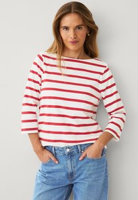 Camisa de rayas rojas y blancas de manga larga con escote barco, hecha de una tela suave, combinada con unos vaqueros azul oscuro de talle alto. Diseño sencillo.