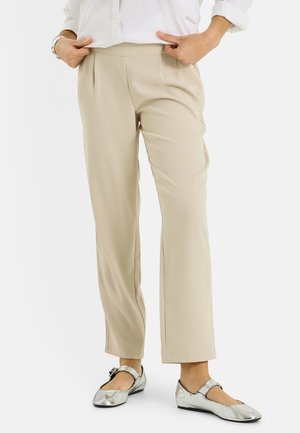 Femme portant un pantalon beige à jambes droites et des ballerines Mary Jane argentées, tenant la taille du pantalon avec les deux mains.