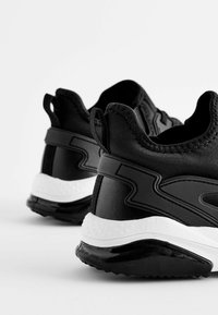 Des chaussures de sport noires avec un dessus en synthétique lisse, une semelle intermédiaire blanche, une semelle extérieure texturée et une languette à l'arrière pour un enfichier facile.