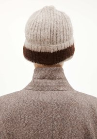 Persoon met een beige en bruine gebreide beanie en een bronzen textuur jas, gezien van achteren tegen een effen achtergrond.