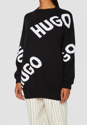 Personne portant un pull noir oversize avec le texte blanc "HUGO" en diagonale sur le devant et les manches, associé à un pantalon blanc à fines rayures.