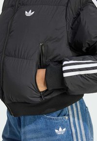 Zwarte pufferjack met zichtbare ritsen, gecombineerd met blauwe denim met drie witte strepen en een Adidas-logo op de zakken.