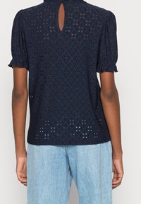 Blusa blu navy con motivo a occhio di diamante, maniche a sbuffo e dettaglio a chiave sulla schiena, realizzata in tessuto leggero. Abbinata a jeans azzurri.