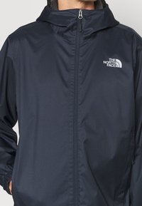 Mann, der eine dunkelblaue, zipbare Kapuzenjacke mit dem Logo von The North Face auf der Brust trägt und die Hände in den Taschen hat.