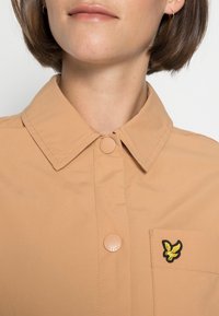 Camicia button-up tan con colletto, dotata di tasca e logo ricamato giallo a forma di uccello. Tessuto liscio con una leggera lucentezza.