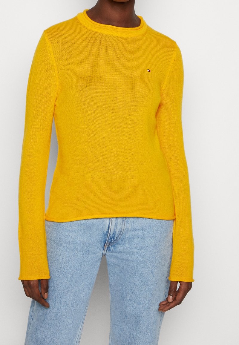 Femme portant un pull jaune vif à manches longues avec un petit logo sur la poitrine et un jean en denim bleu clair, debout contre un mur blanc.