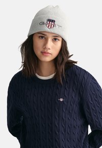 Grau gerippte Strickmütze mit einem Logo-Patch, kombiniert mit einem dunkelblauen Kabelstrickpullover, der ein kleines Logo auf der Brust trägt.