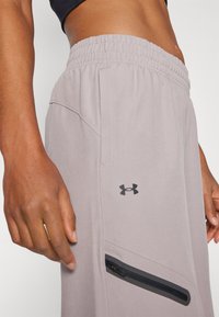 Vaaleanharmaat urheiluhousut, joissa on istuva vyötärönauha, sivutasku mustalla vetoketjulla ja musta Under Armour -logo reiden kohdalla.
