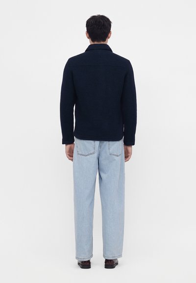 Homme debout, dos tourné, portant une veste sombre, un jean bleu clair et des chaussures foncées sur un fond blanc uni.
