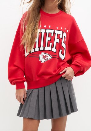Osoba nosząca czerwoną bluzę Kansas City Chiefs na szarej plisowanej spódnicy, stojąca z jedną ręką poprawiającą dół bluzy.