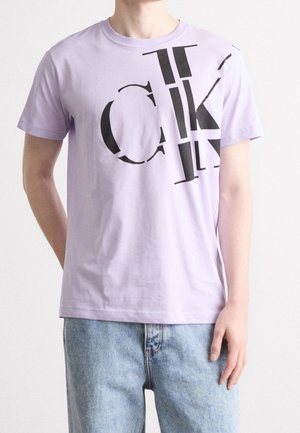 T-shirt lilas à manches courtes en coton, avec des lettres graphiques noires en gras et un col rond classique. Porté avec un jean bleu clair.