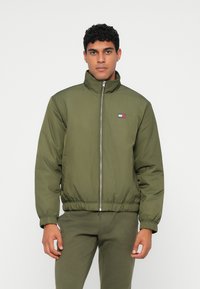 Ελαιοπράσινο μπουφάν με επένδυση, υψηλό γιακά, μπροστινό φερμουάρ και ελαστικές μανσέτες. Διαθέτει ένα μικρό λογότυπο σε κόκκινο, λευκό και μπλε χρώμα.