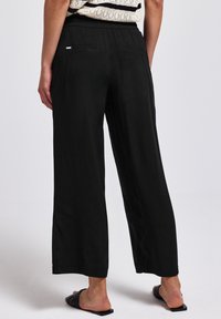 Pantalons noirs à jambes larges avec une taille élastique, dotés de deux poches arrière. Texture lisse, coupés juste au-dessus de la cheville.