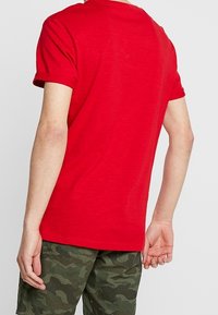 Rød kortærmet t-shirt med rund hals, lavet af blødt stof. Tætsiddende stil, afslutning ved hoften, båret med shorts med camouflage-mønster.
