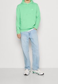 Felpa in cotone verde menta con tasca frontale e cappuccio con coulisse, abbinata a jeans blu chiaro e sneakers bianche con accenti colorati.