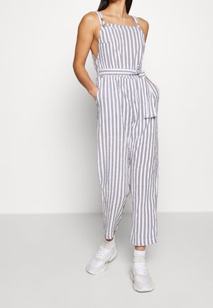 Combinaison à rayures gris et blanc, dotée de bretelles ajustables, d'une ceinture à nouer à la taille et de poches latérales. Portée avec des baskets blanches.