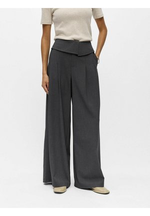 Personne portant un pantalon taille haute à jambes larges de couleur charbon avec une ceinture repliée, un haut beige et des chaussures crème, debout devant un fond uni.