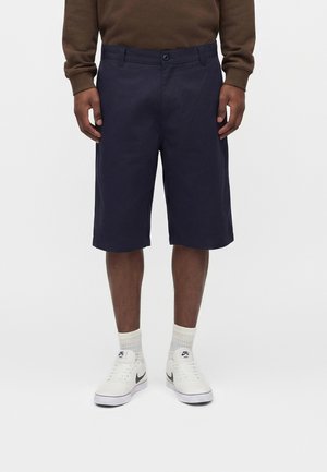 Mężczyzna ubrany w granatowe spodenki do kolan, brązową koszulę z długim rękawem, białe sneakersy marki Nike i białe skarpety, stojący na jednolitym tle.